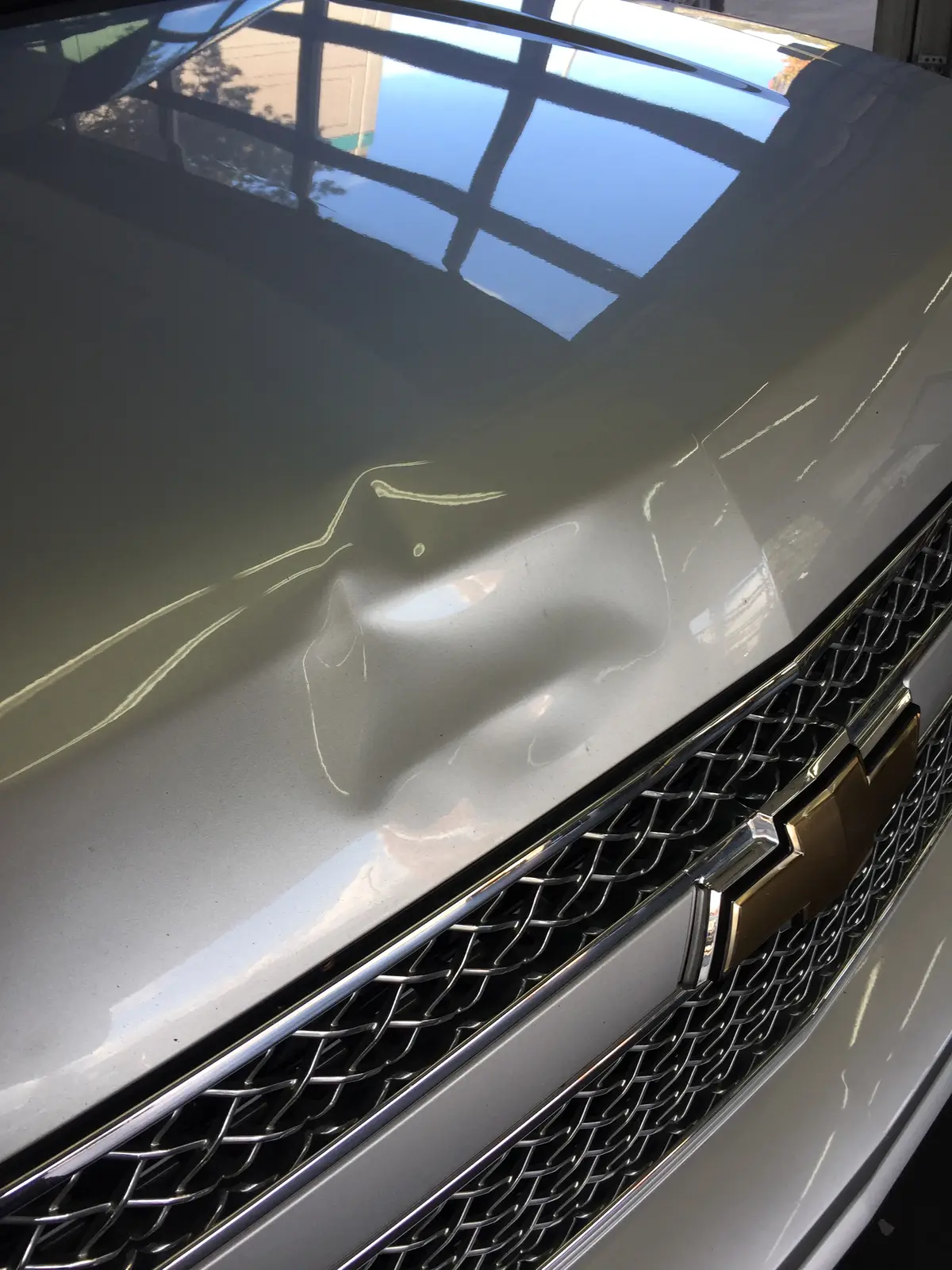 A dent on a silver Chevrolet hood just above the grille, before repair