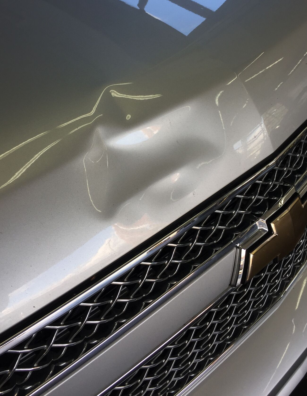 A dent on the hood of a silver Chevrolet above the grille, before paintless dent repair in Bellevue